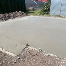 Michał Szymczuk - Świeżo wylana, gładka betonowa płyta fundamentowa z drewnianym szalunkiem, widoczny pryzmat ziemi i zielone tuje w tle.