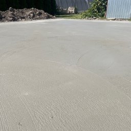 Michał Szymczuk - Świeżo wylana, gładka betonowa posadzka zewnętrzna z widocznymi śladami po wygładzaniu, z pryzmą ziemi i fragmentem ogrodzenia w tle.