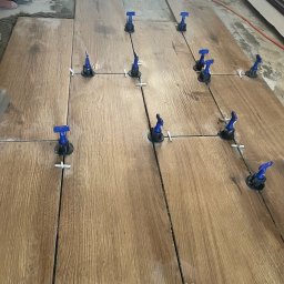 Michał Szymczuk - Układanie płytek podłogowych imitujących drewno z użyciem systemu poziomowania z niebieskimi klinami i białymi krzyżykami dystansowymi.