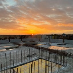 Michał Szymczuk - Wylany strop budynku w trakcie budowy, z widocznym zbrojeniem i szalunkiem, na tle pomarańczowego zachodu słońca i zabudowy miejskiej.