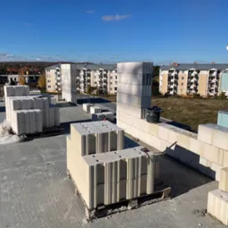 Wznoszenie ściany z bloczków silikatowych na budowie, palety z materiałem i częściowo wymurowana ściana na tle zabudowy wielorodzinnej w słoneczny dzień.