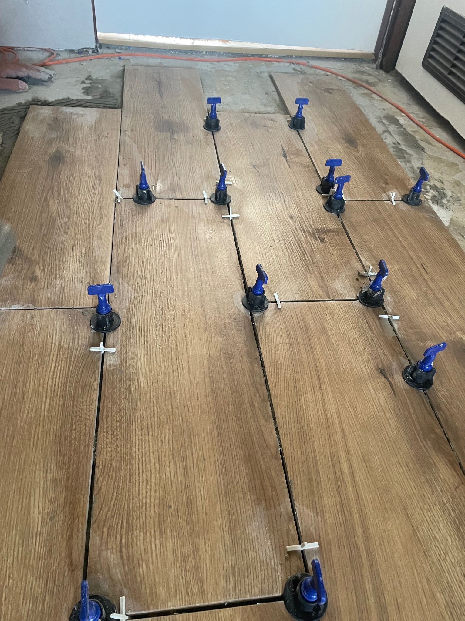 Układanie płytek podłogowych imitujących drewno z użyciem systemu poziomowania z niebieskimi klinami i białymi krzyżykami dystansowymi.