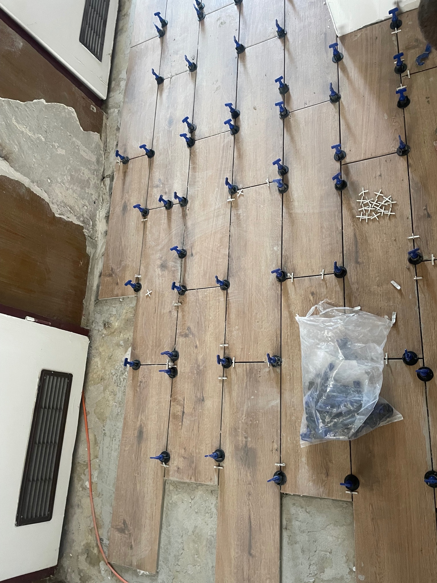 Układanie podłogi z płytek imitujących drewno, z użyciem systemów poziomowania płytek, widoczna surowa ściana w tle.