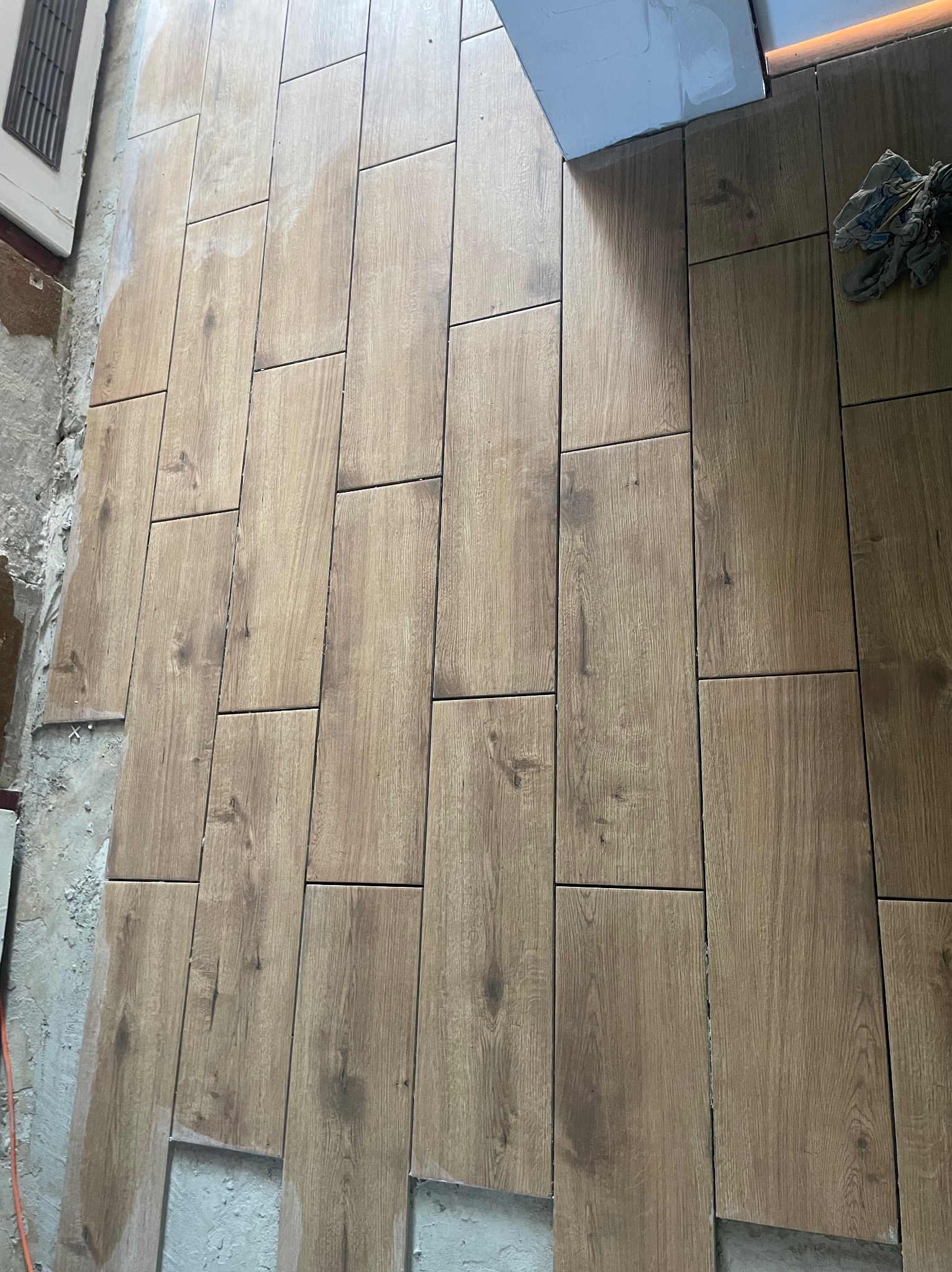 Układanie podłogi z płytek imitujących drewno, widoczne niedokończone krawędzie i zaprawa, narzędzie w rogu.