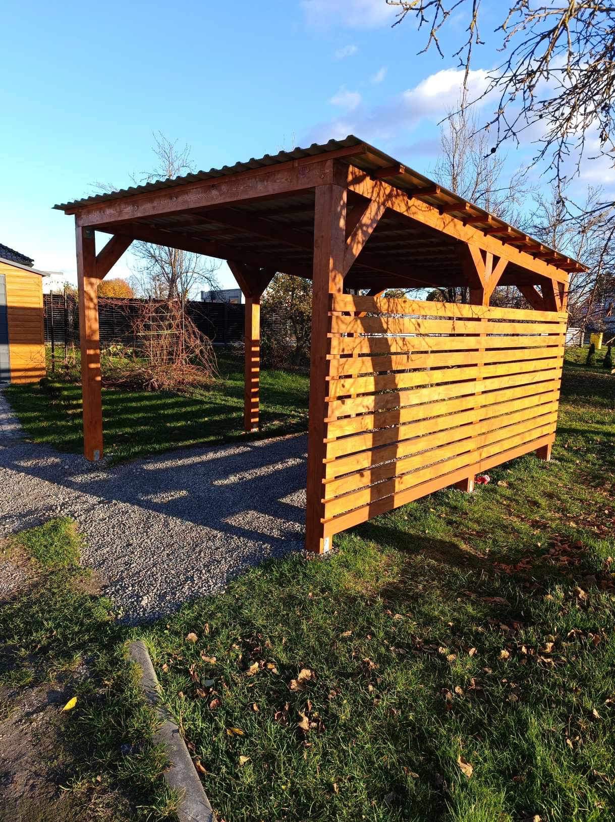 Drewniana wiata samochodowa z częściową osłoną z desek, zadaszona blachą falistą, na trawniku z kamiennym podjazdem, w tle budynek gospodarczy i drzewa.