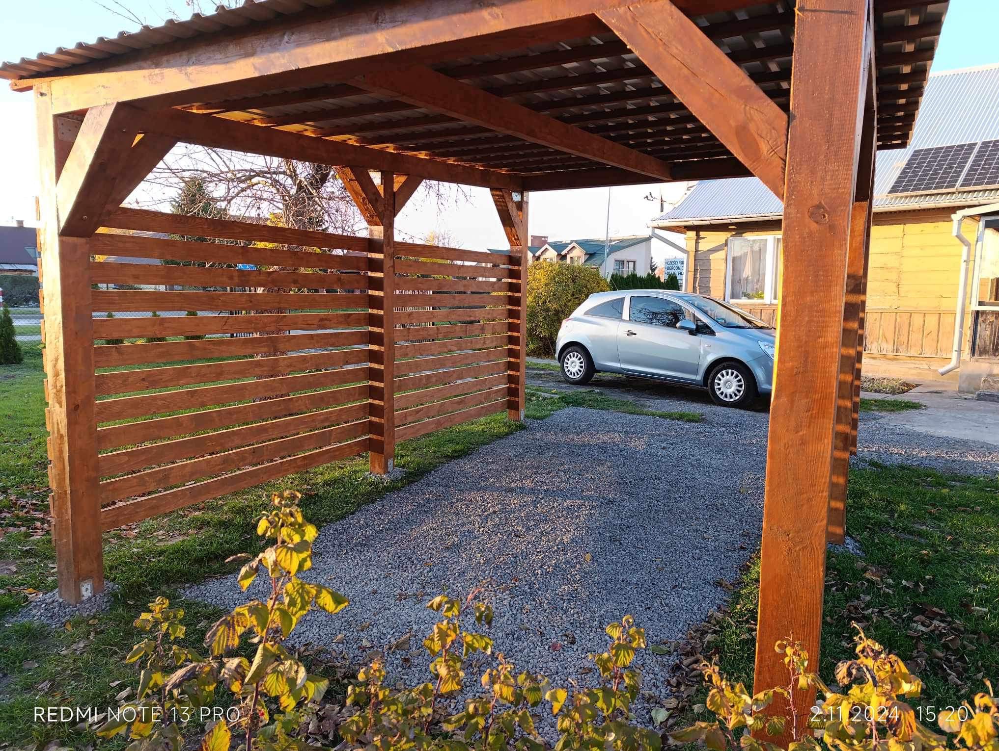 Drewniana wiata garażowa z ażurową ścianą boczną, pokryta blachą trapezową, na żwirowym podłożu, z zaparkowanym srebrnym samochodem osobowym w tle.