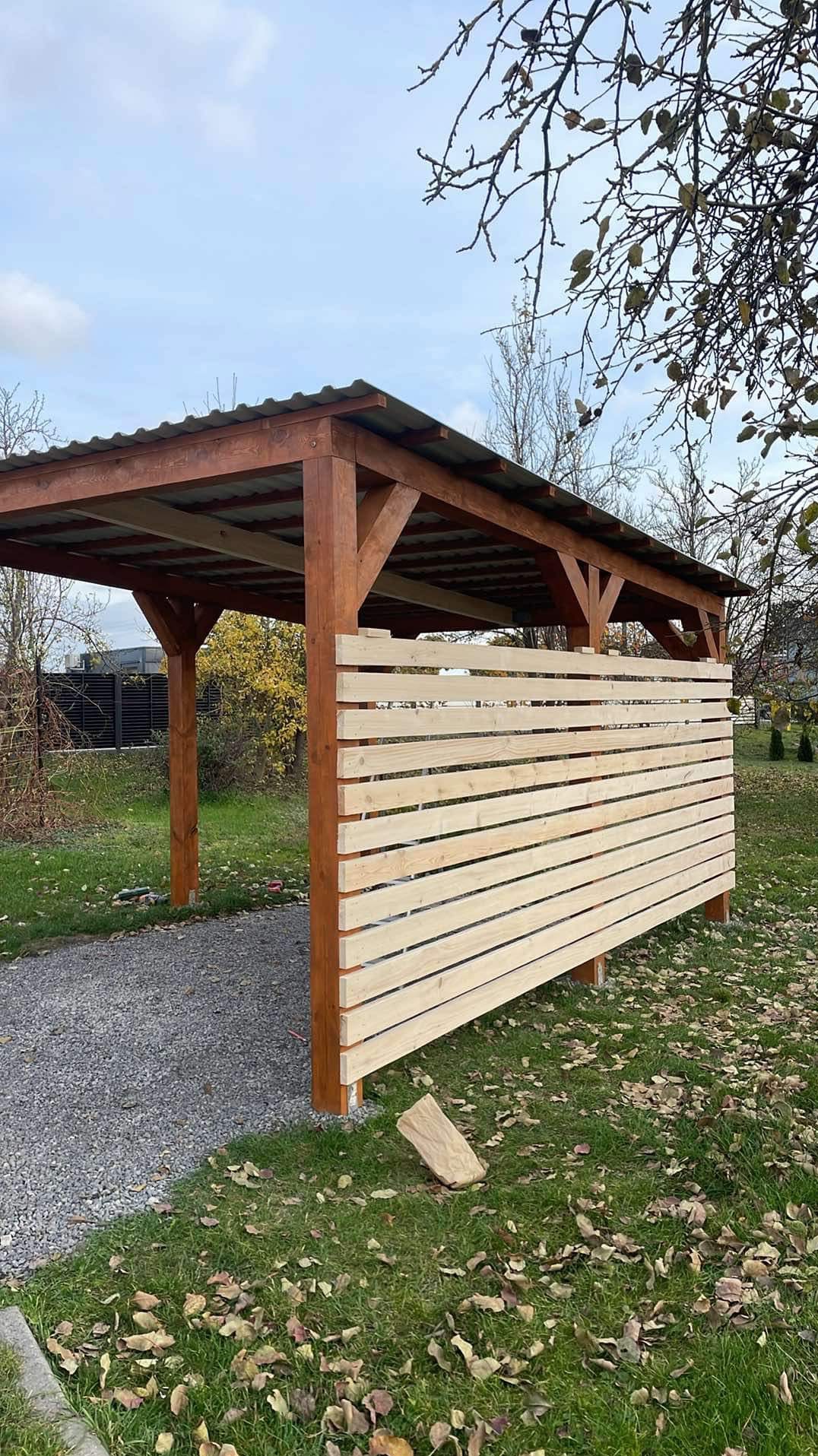 Drewniana wiata z częściowo zabudowaną ścianą z poziomych desek, widok z zewnątrz, na trawie rozsypane liście.