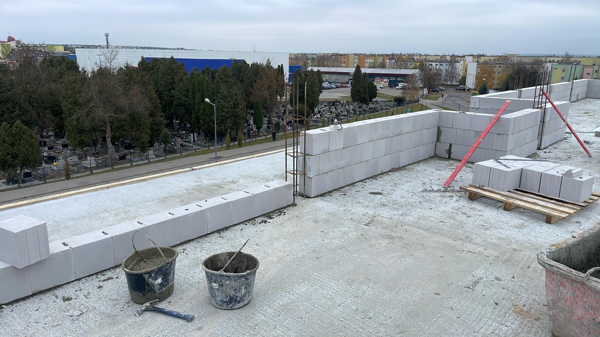 Wznoszenie ściany z bloczków betonowych na dachu, widoczne zbrojenie, poziomica i paleta z bloczkami, w tle cmentarz i zabudowa miejska.