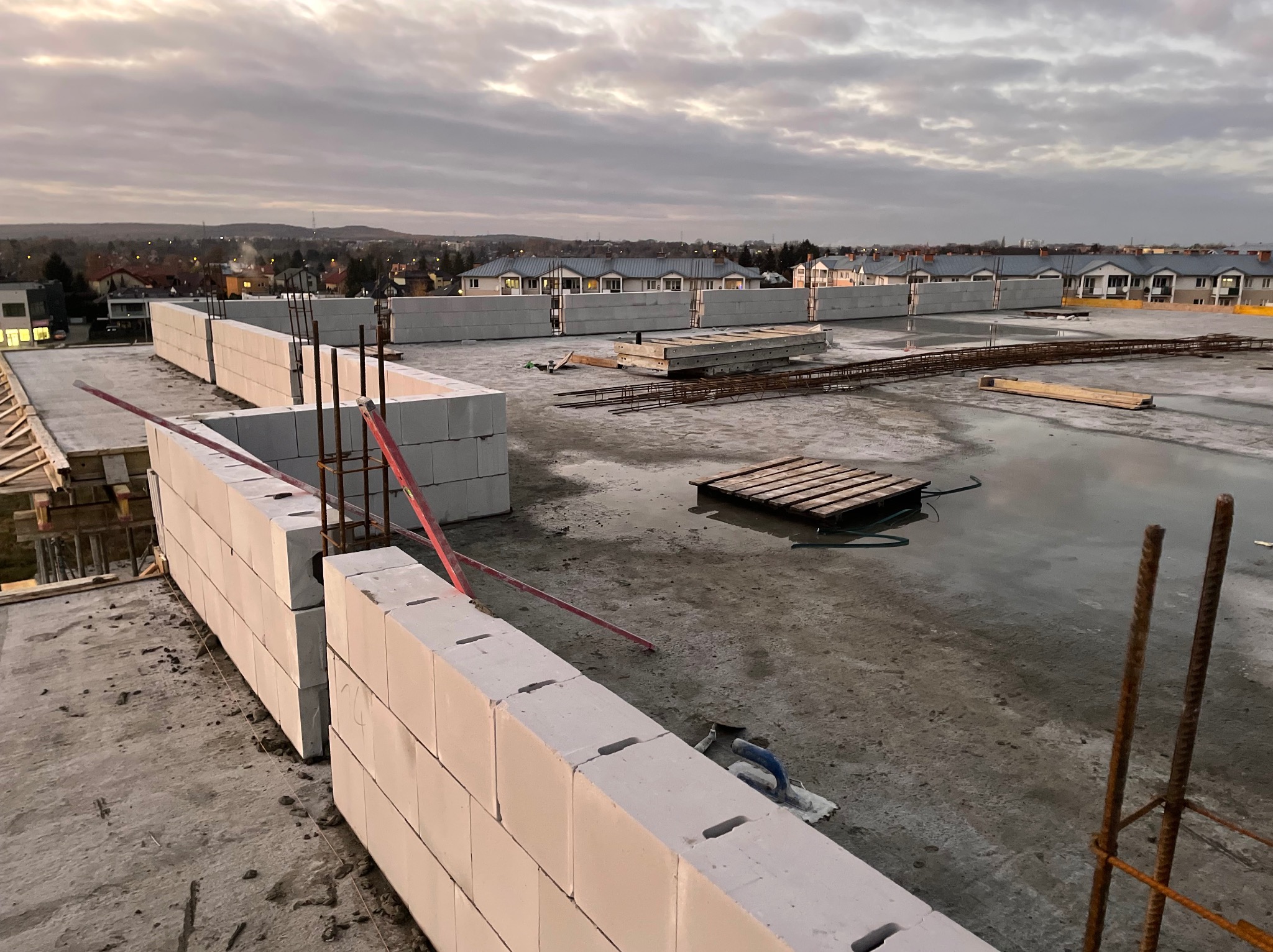 Wznoszenie muru z betonu komórkowego na mokrym stropie budynku wielorodzinnego, widoczne zbrojenia i poziomica, w tle zabudowa osiedlowa pod pochmurnym niebem.
