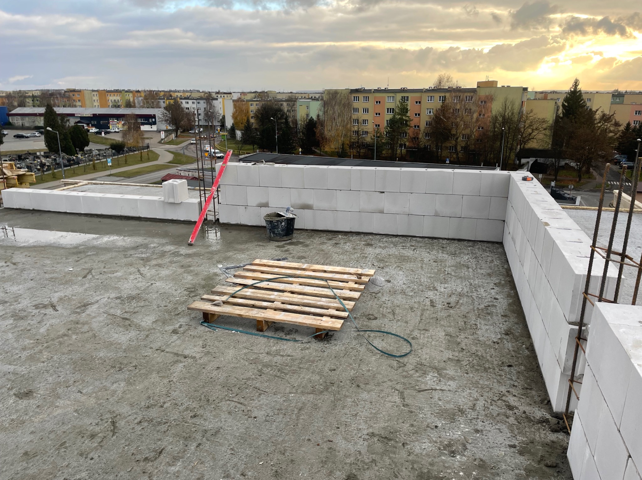 Wznoszenie ścian z białych bloczków na betonowym stropie, widoczna paleta, wiadro i poziomica, w tle zabudowa miejska pod pochmurnym niebem.