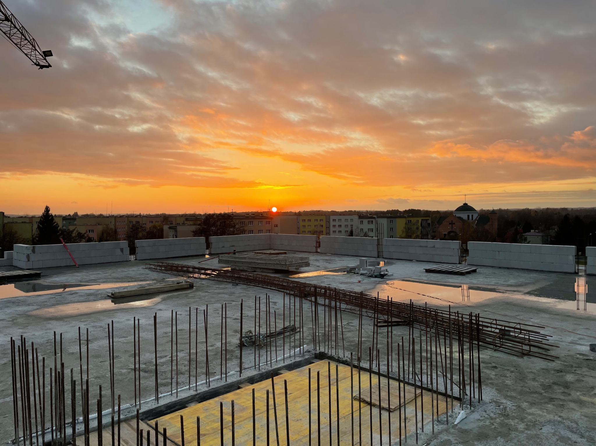 Wylany strop budynku w trakcie budowy, z widocznym zbrojeniem i szalunkiem, na tle pomarańczowego zachodu słońca i zabudowy miejskiej.