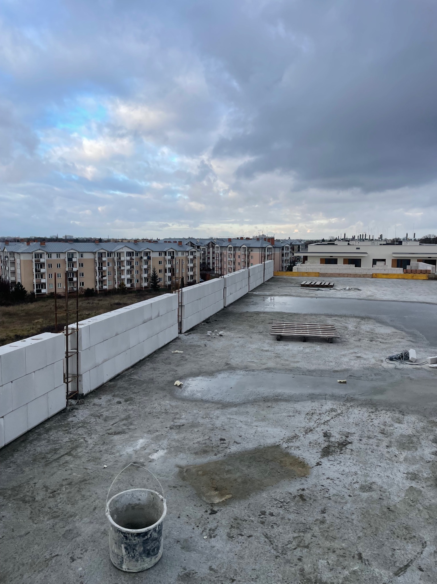 Wznoszenie ściany z białych bloczków na mokrym, betonowym stropie w trakcie budowy domu, widoczne zbrojenie i wiadro, w tle budynki mieszkalne pod pochmurnym niebem.