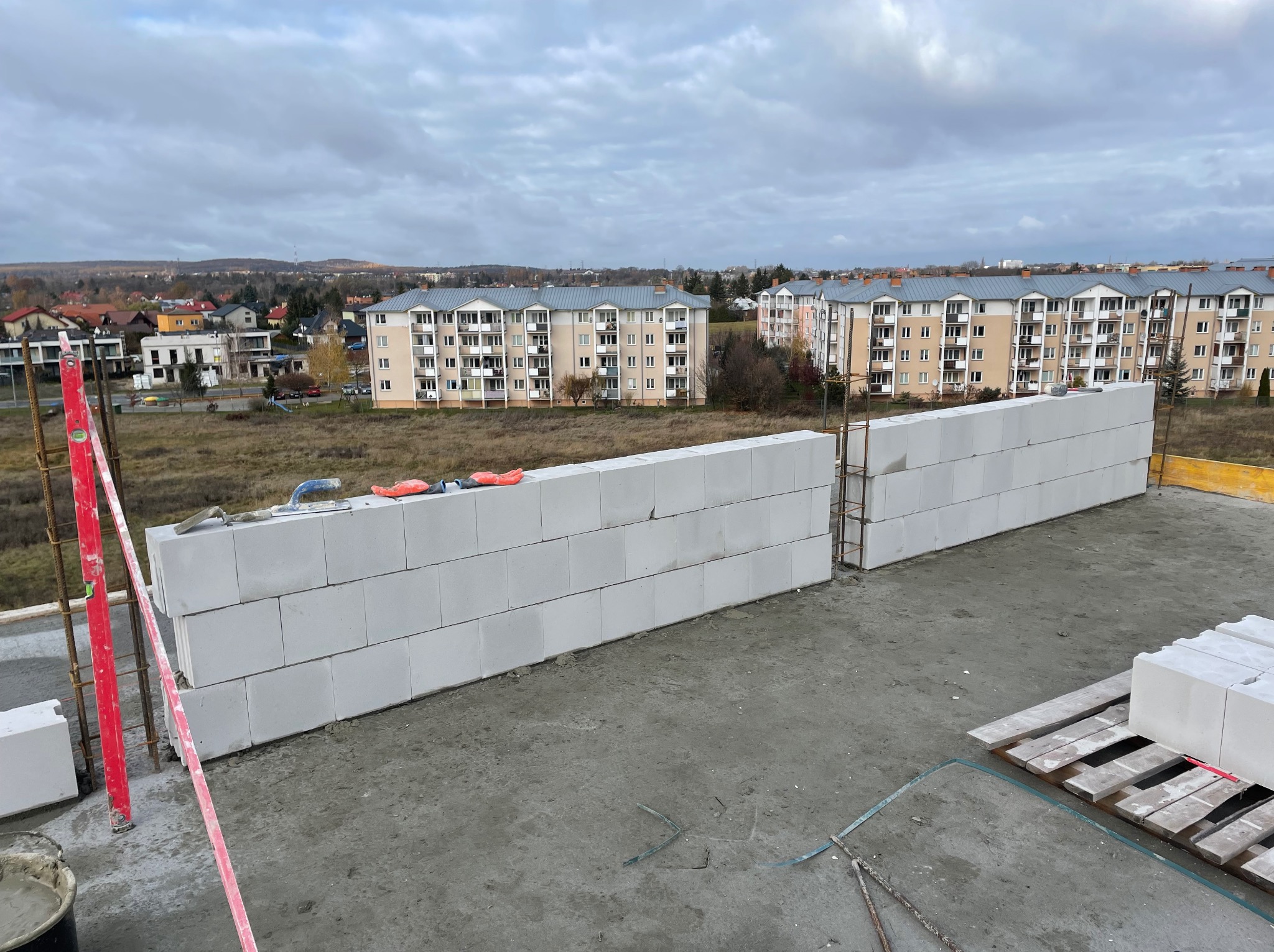 Wznoszenie ściany z białych bloczków betonu komórkowego Ytong na tle zabudowy wielorodzinnej i pochmurnego nieba, widoczny poziomica, zaprawa i narzędzia murarskie.