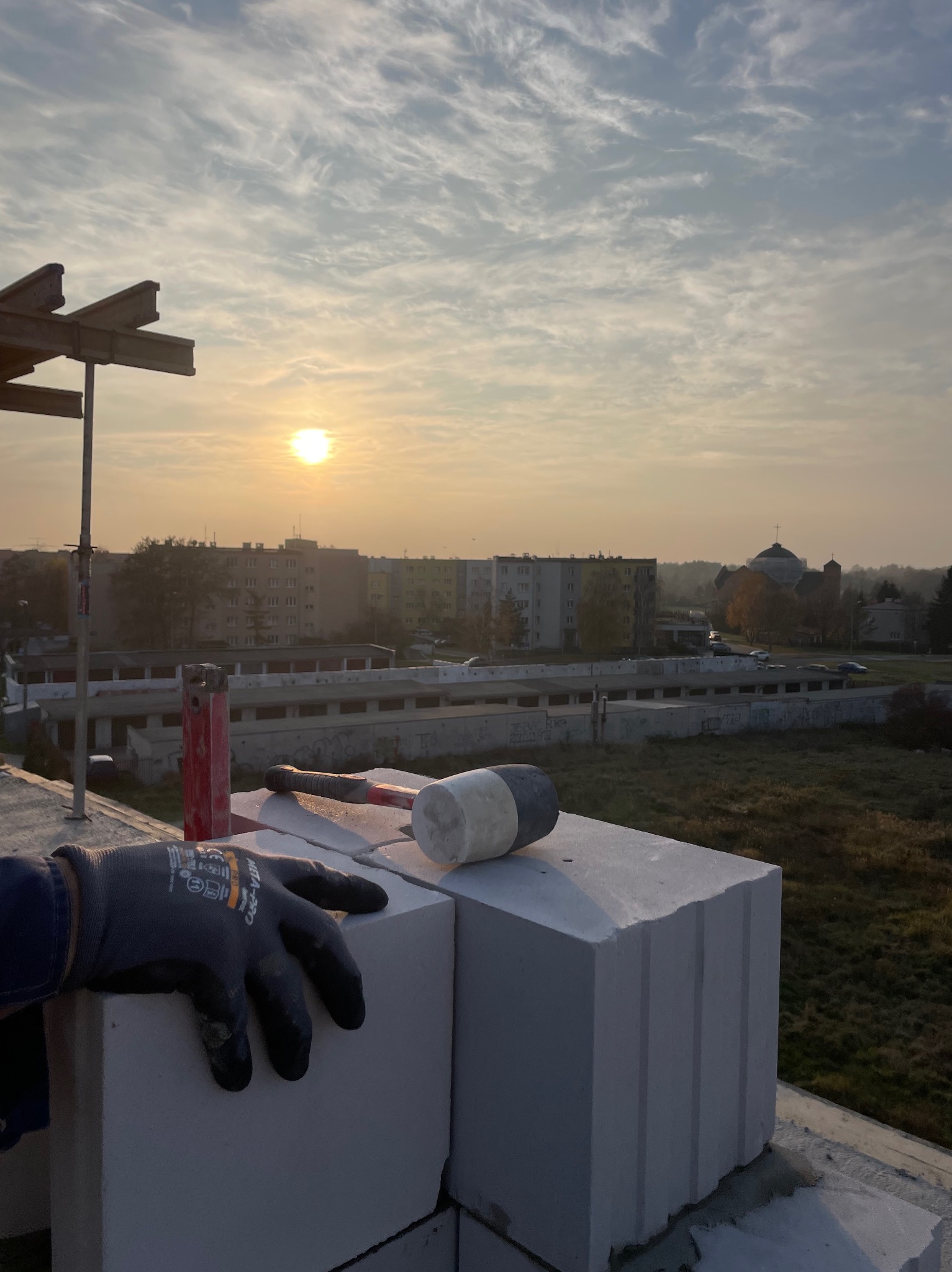 Wznoszenie ściany z białych bloczków, widok z góry na murarza w rękawicy, gumowy młotek i poziomica w tle zabudowa miejska i zachodzące słońce.