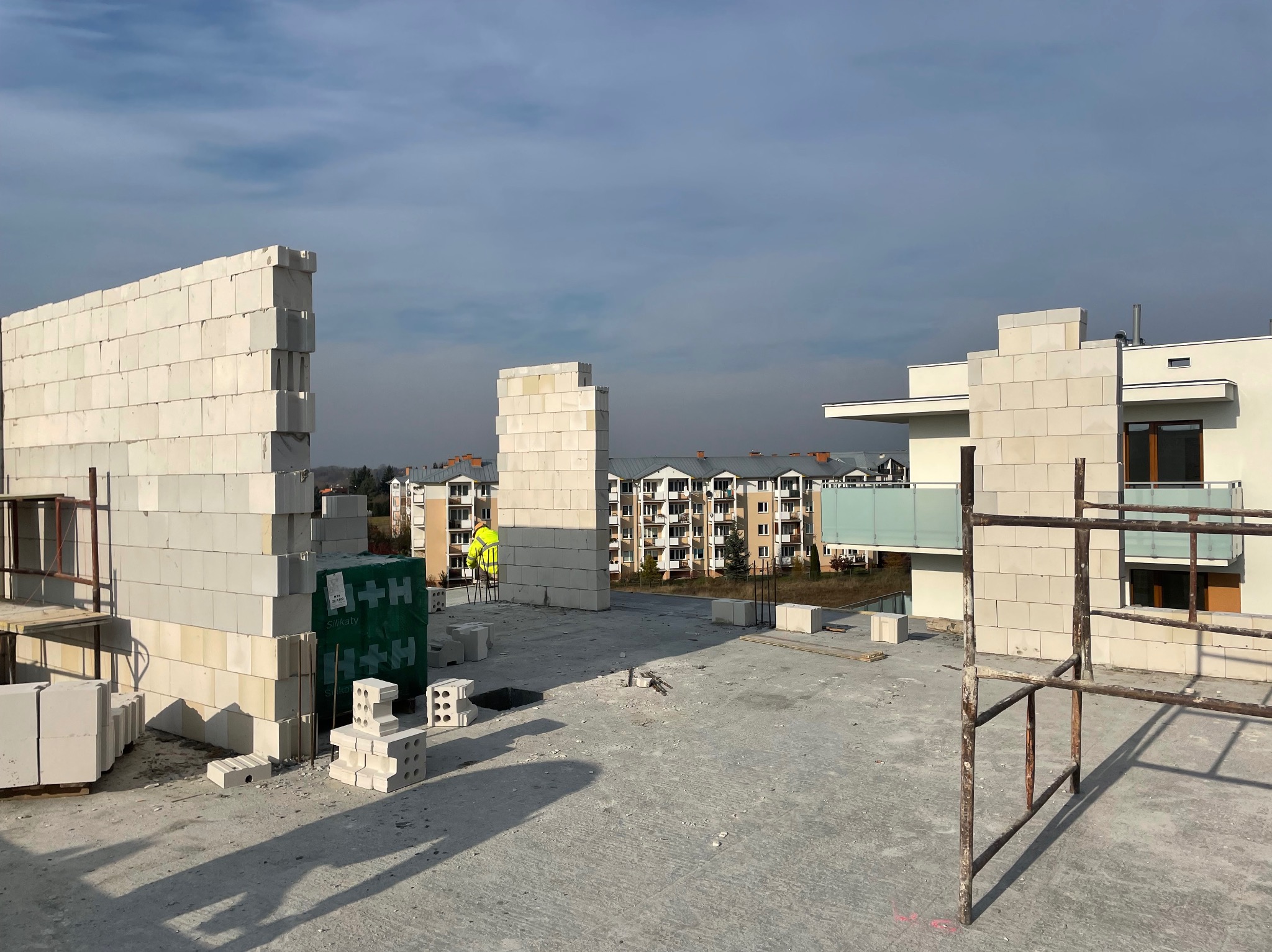 Wznoszone ściany z białych bloczków silikatowych na dachu budynku wielorodzinnego, widoczny pracownik w żółtej kamizelce oraz rusztowanie, w tle zabudowa miejska.