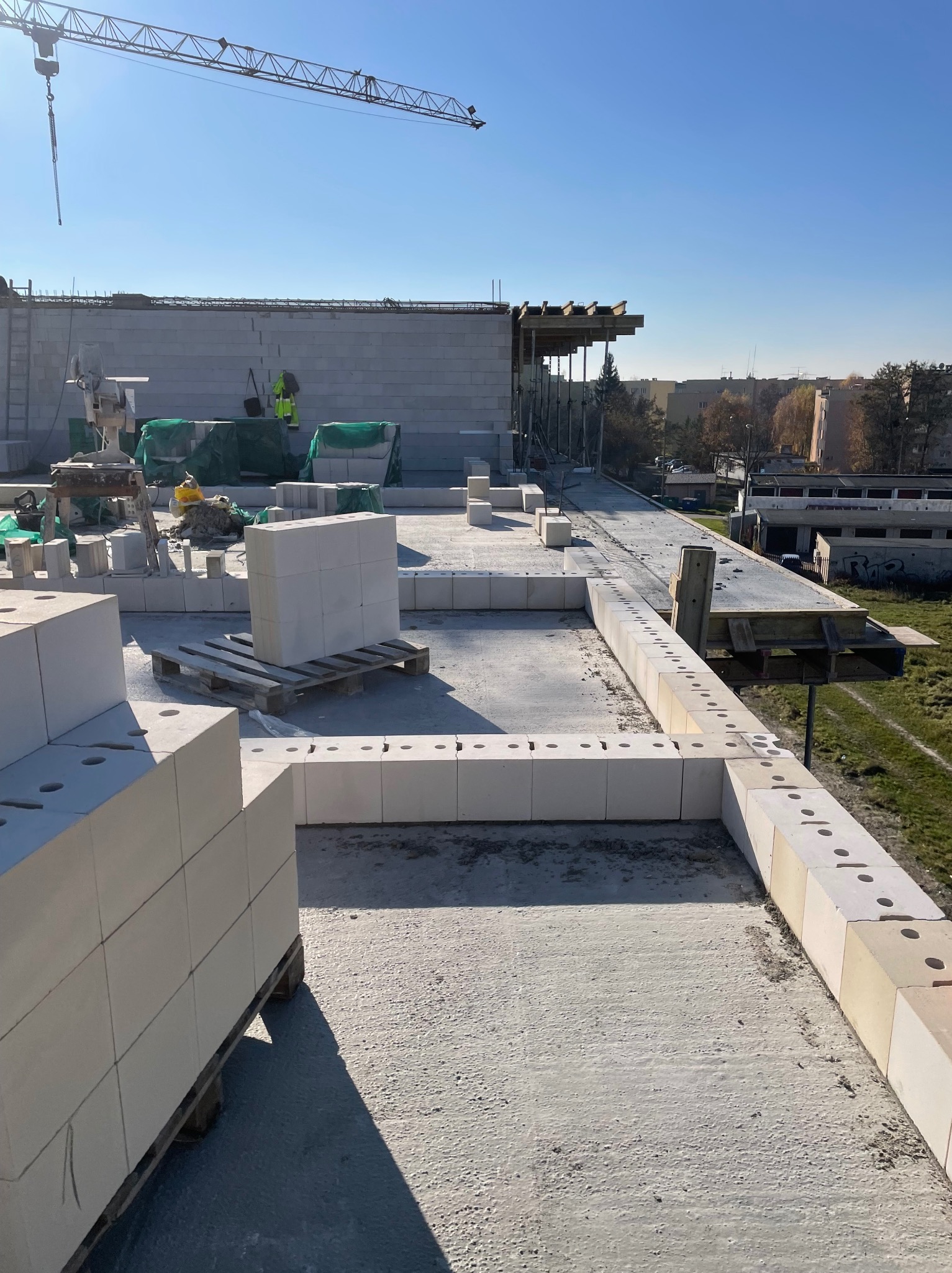 Wznoszenie ścian z bloczków betonu komórkowego na budowie, widok z góry, paleta z bloczkami w centrum kadru, w tle częściowo widoczny żuraw budowlany.