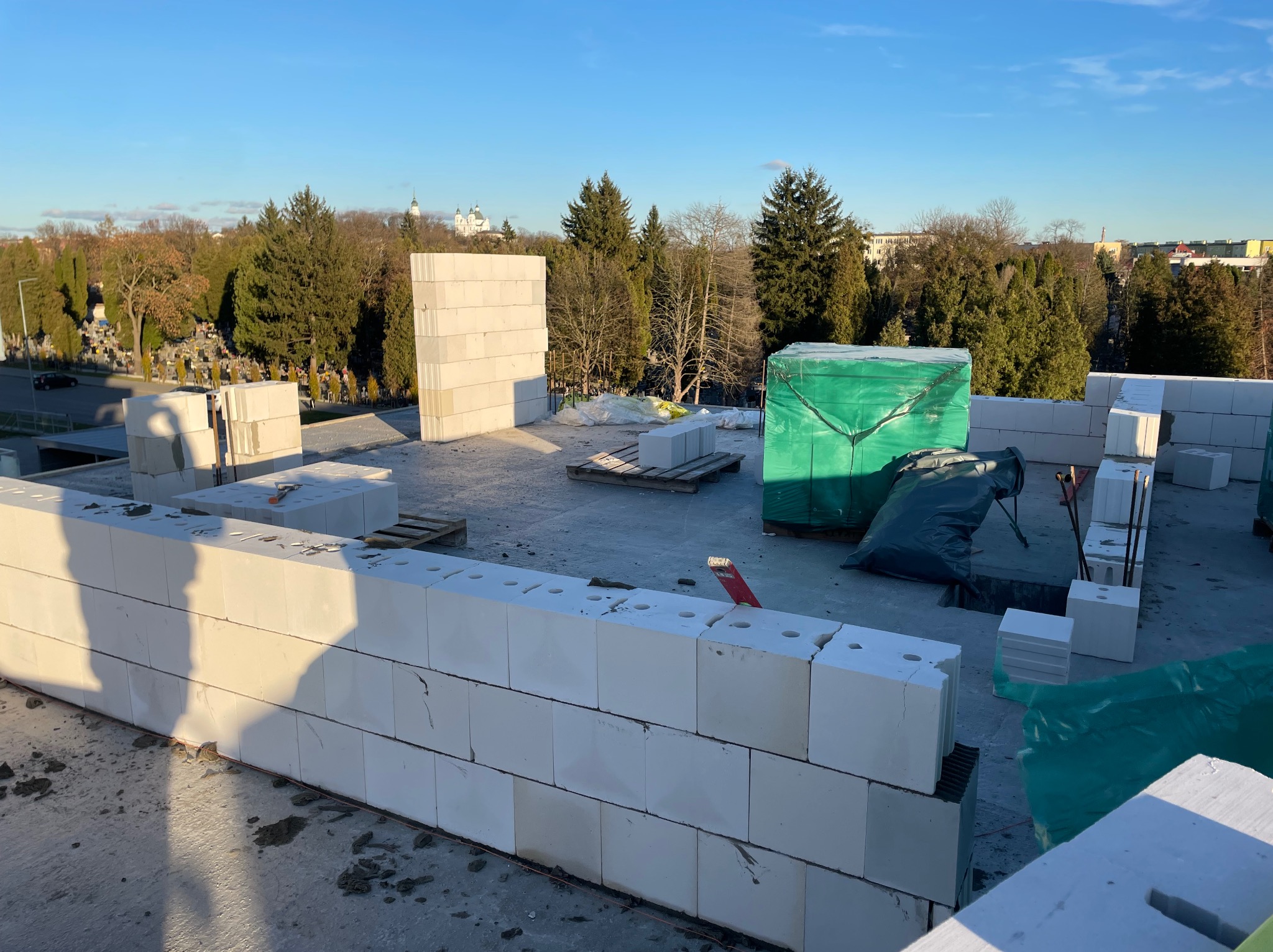 Wznoszenie ścian z betonu komórkowego Ytong na budowie domu, widok z góry na mur z widocznymi zapasami materiału budowlanego i odległą panoramą miasta z zielenią i kościołem.