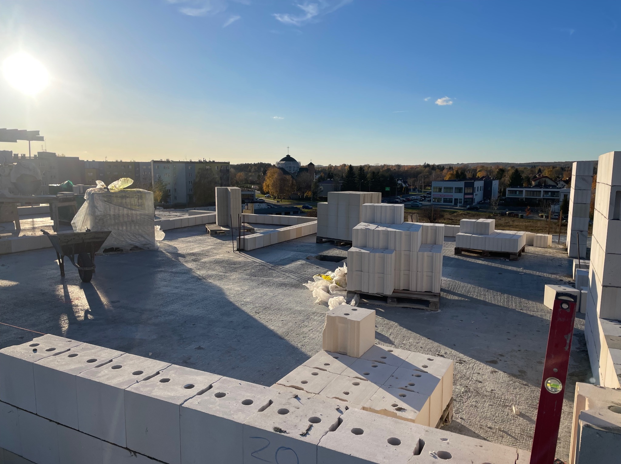 Wznoszenie ścian domu z betonu komórkowego na tle zabudowy miejskiej w słoneczny dzień, widoczny poziomica i taczka.