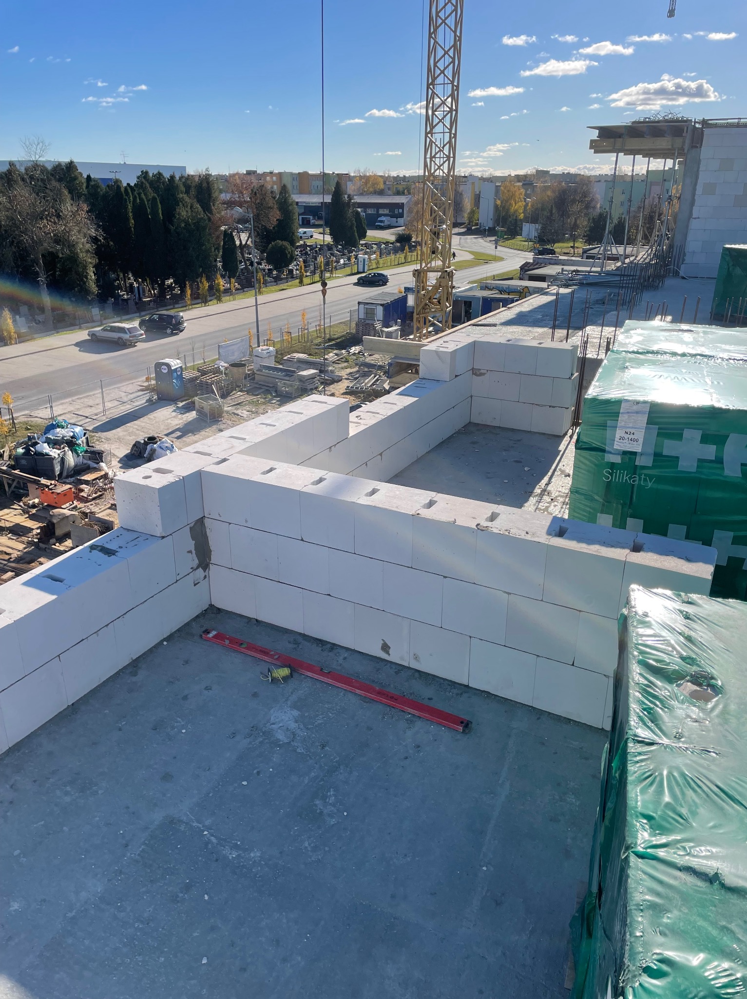 Wznoszenie ścian parteru z białych bloków silikatowych na budowie domu, widoczny poziomica murarska i paleta zafoliowanych bloków, w tle ulica z ruchem samochodowym i budynki.