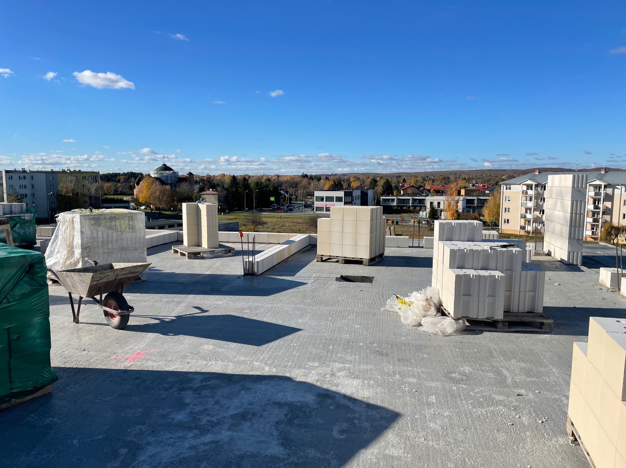 Wysoki kąt widoku na plac budowy z bloczkami ceramicznymi na paletach, częściowo wymurowanymi ścianami i taczką, w tle budynki mieszkalne i błękitne niebo.