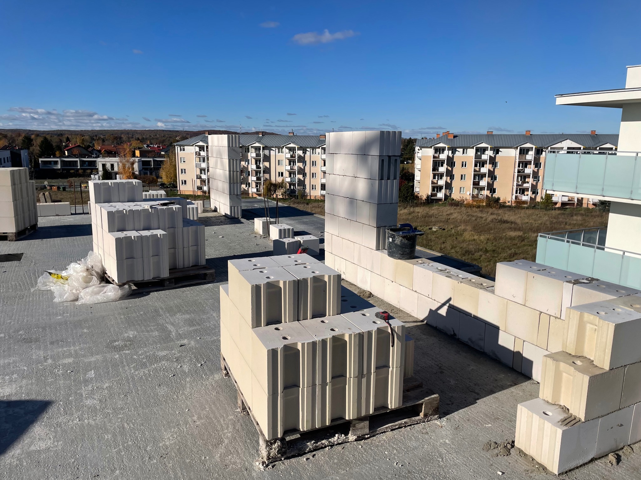 Wznoszenie ściany z bloczków silikatowych na budowie, palety z materiałem i częściowo wymurowana ściana na tle zabudowy wielorodzinnej w słoneczny dzień.