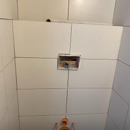KeramiCraft Robert Rant - Białe płytki ścienne w łazience z widoczną instalacją podtynkową i rurami odpływowymi. Etap prac wykończeniowych, widok z dołu.