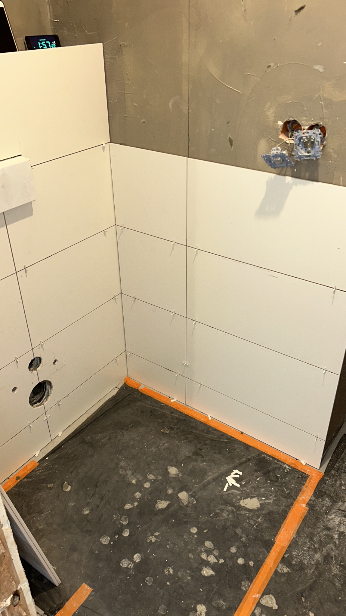 Świeżo ułożone, białe płytki ścienne w łazience, z widocznymi krzyżykami dystansowymi i przygotowanym miejscem na armaturę. Podłoga zabezpieczona folią.