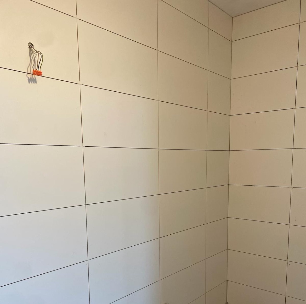 Wykończona ściana w łazience z białymi, prostokątnymi płytkami. Widoczne przewody elektryczne wystające ze ściany. Minimalistyczny design.