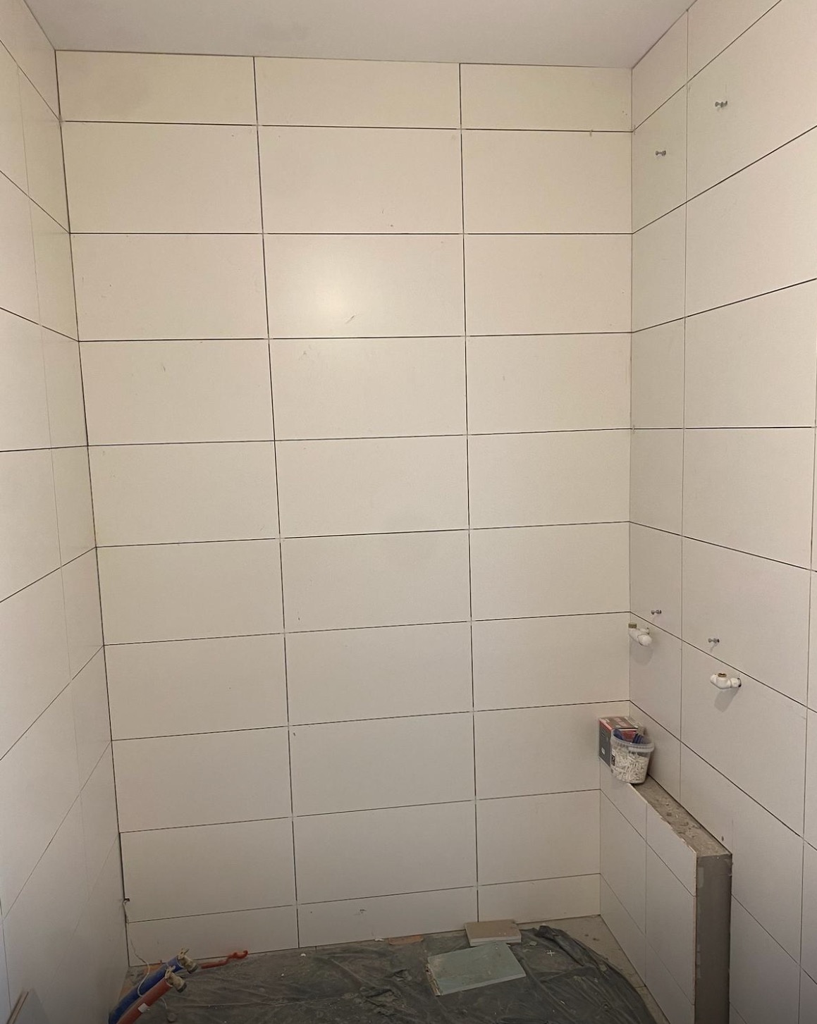 Wykończona ściana łazienki z białymi, prostokątnymi płytkami. Widoczne przygotowane punkty instalacji wodnej i kanalizacyjnej. Na podłodze folia ochronna i resztki materiałów.