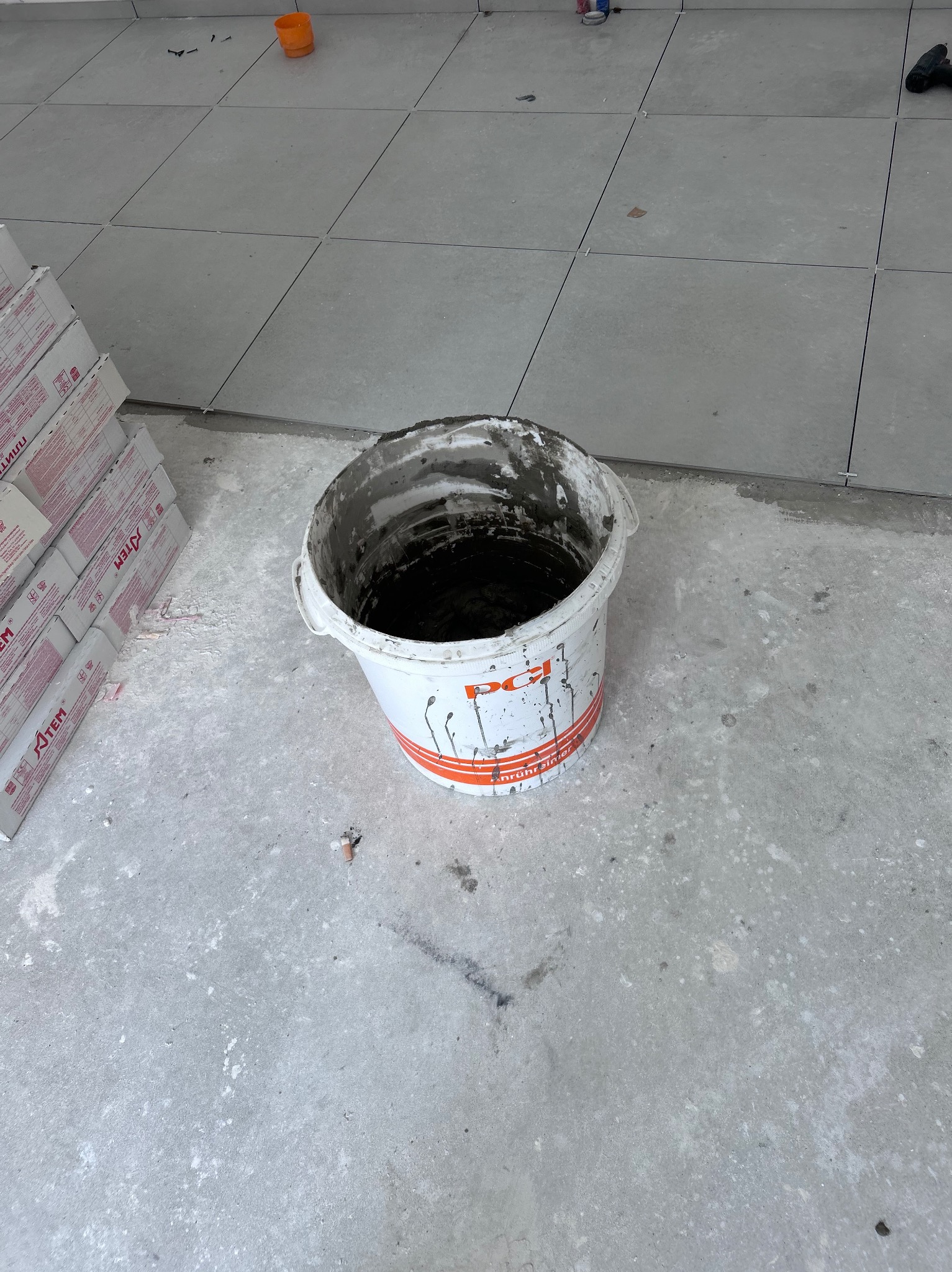 Wiadro z zaprawą na tle ułożonych, szarych płytek podłogowych w Lublińcu. Widoczne paczki płytek i narzędzia. Prace glazurnicze w toku, etap przygotowania zaprawy.