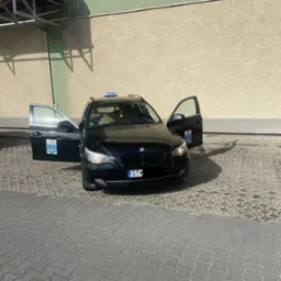 Czarny samochód kombi z otwartymi drzwiami, oznaczony jako taksówka, zaparkowany na brukowanym placu przed beżowym budynkiem z zielonym akcentem.