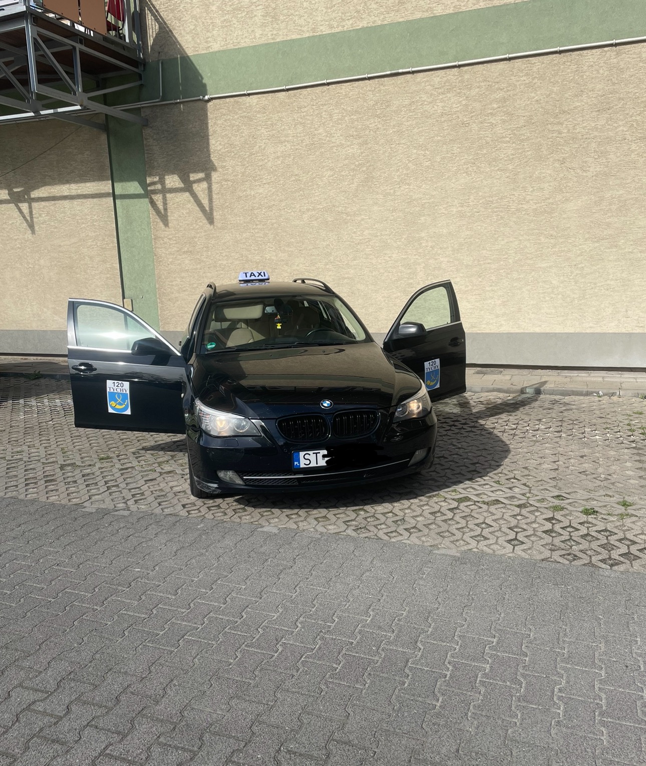 Czarny samochód kombi z otwartymi drzwiami, oznaczony jako taksówka, zaparkowany na brukowanym placu przed beżowym budynkiem z zielonym akcentem.