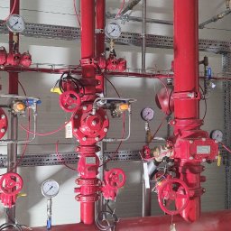 ABM Service plus - Czerwone rury instalacji przeciwpożarowej z manometrami i zaworami, widoczne drabiny w tle.