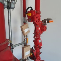 ABM Service plus - Czerwona instalacja przeciwpożarowa z zaworami, rurami i osprzętem, widoczne etykiety na elementach instalacji.