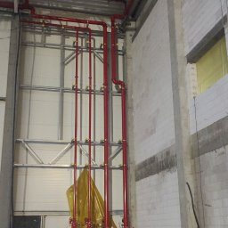 ABM Service plus - Czerwona instalacja rur przeciwpożarowych w surowym wnętrzu hali, z widocznymi elementami mocującymi i żółtą folią ochronną na dole.