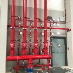 ABM Service plus - Czerwona instalacja przeciwpożarowa z rurami, zaworami i manometrami w hali przemysłowej, widoczne mokre plamy na betonowej posadzce.