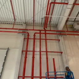 Czerwone rury instalacji przeciwpożarowej zamontowane pod sufitem hali przemysłowej, widoczne mocowania i pracownik na podnośniku koszowym.