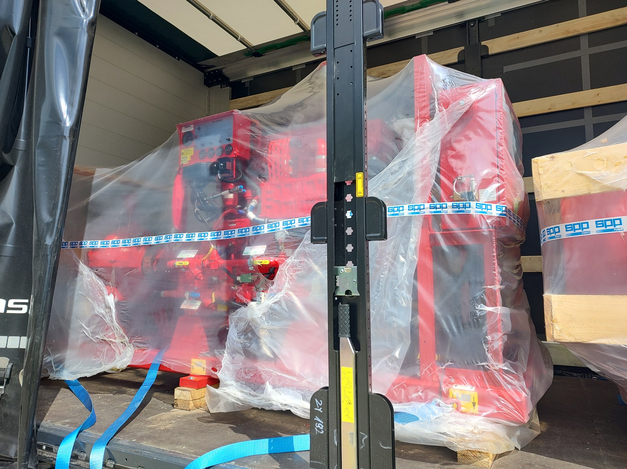 Czerwona pompa hydrauliczna marki Clarke, zabezpieczona folią i taśmą SPP, transportowana wewnątrz ciężarówki, widoczny element wózka widłowego.