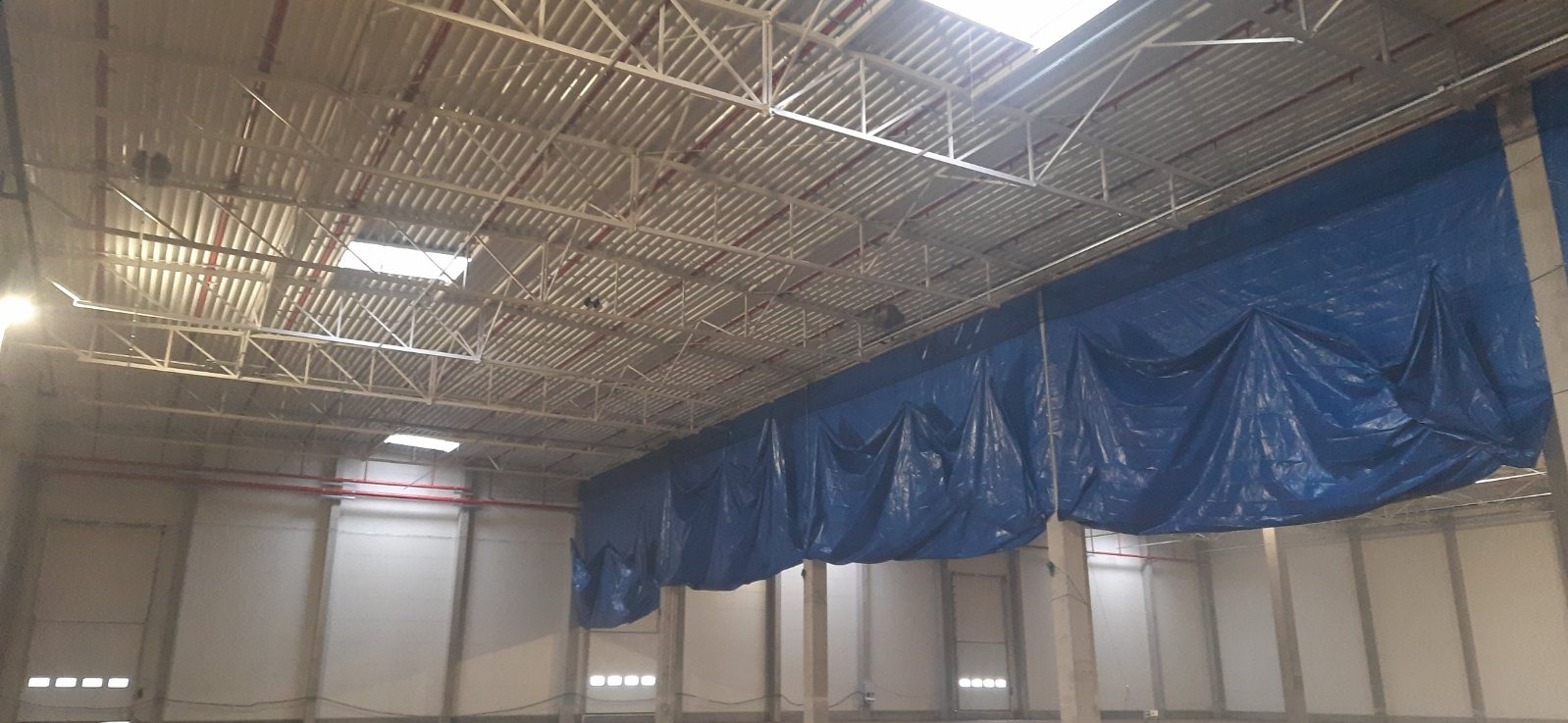 Wnętrze hali magazynowej z widoczną konstrukcją dachu i instalacją tryskaczową pomalowaną na czerwono, zabezpieczoną folią budowlaną przed pracami remontowymi.