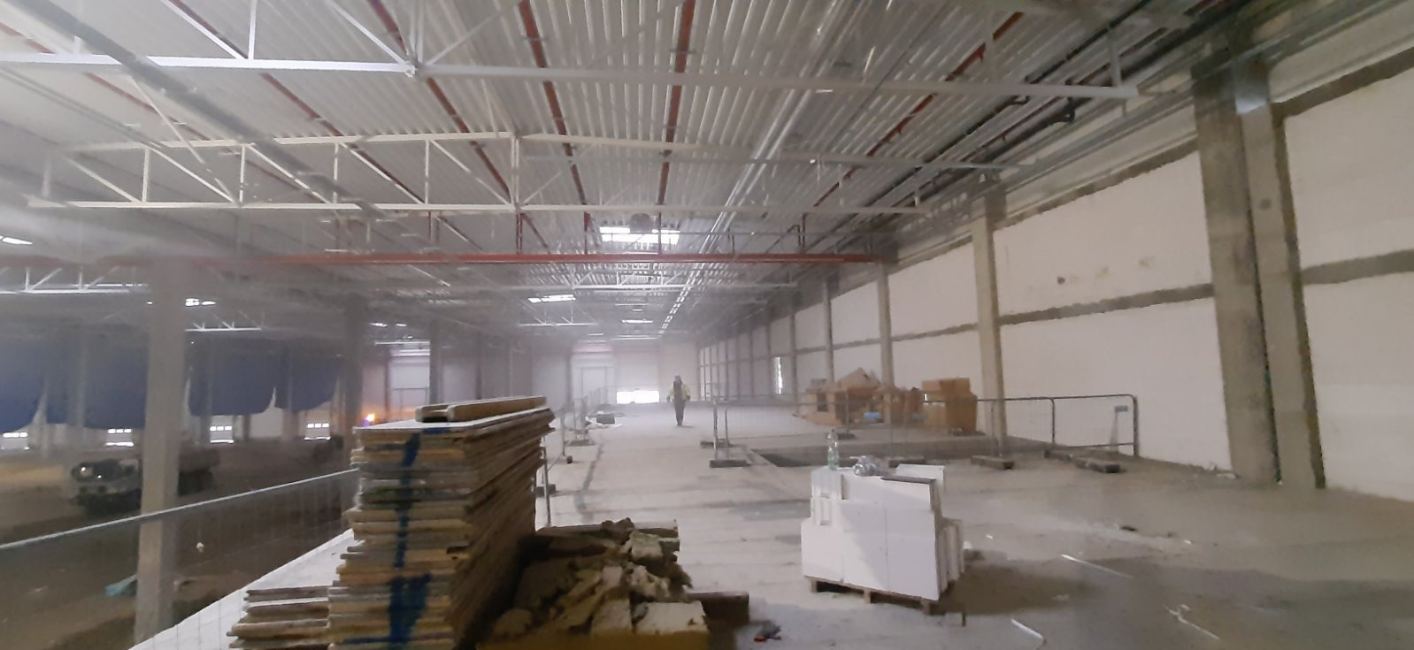 Wnętrze hali przemysłowej w trakcie budowy, widoczne elementy instalacji pod sufitem, stosy materiałów budowlanych i pracownik w oddali.