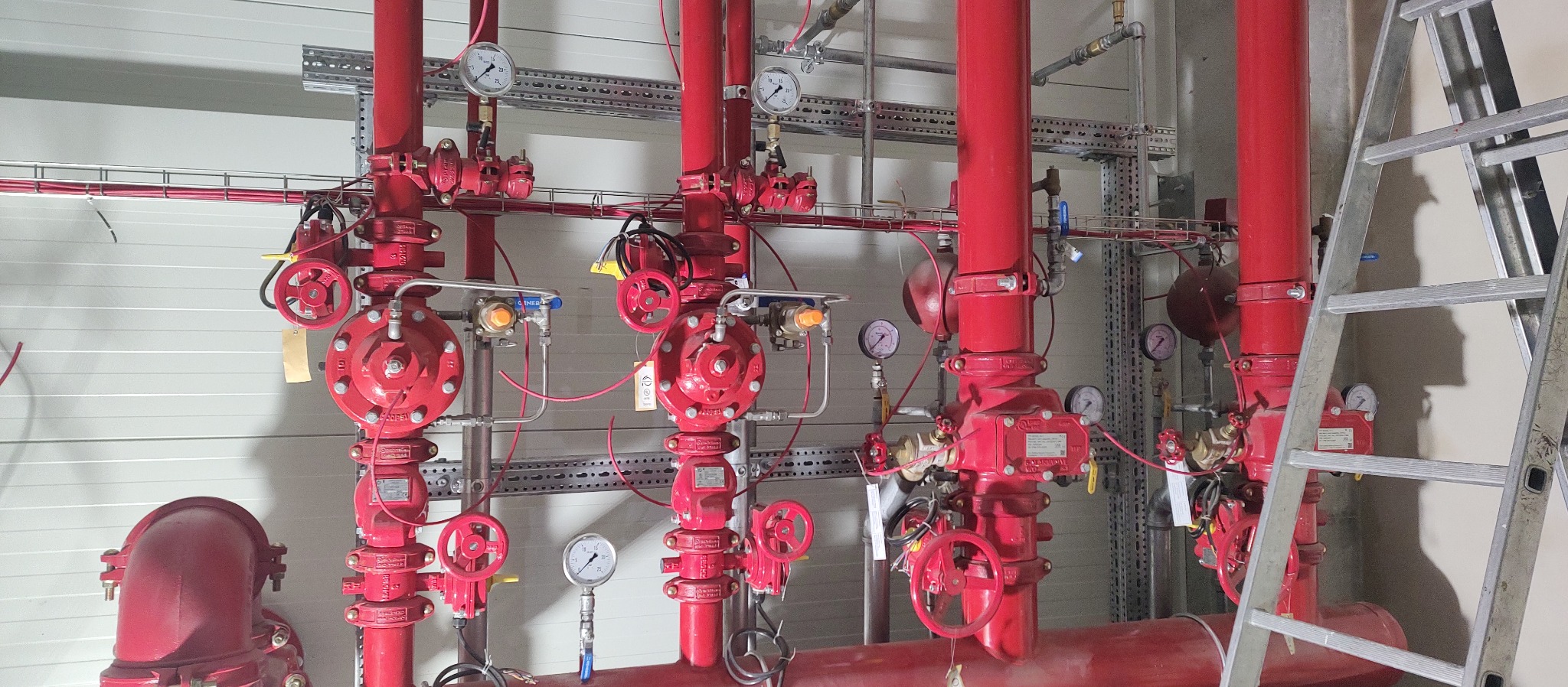 Czerwone rury instalacji przeciwpożarowej z manometrami i zaworami, widoczne drabiny w tle.