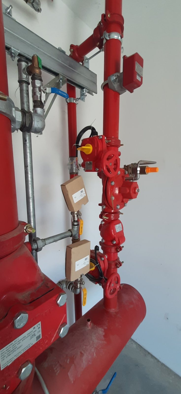 Czerwona instalacja przeciwpożarowa z zaworami, rurami i osprzętem, widoczne etykiety na elementach instalacji.