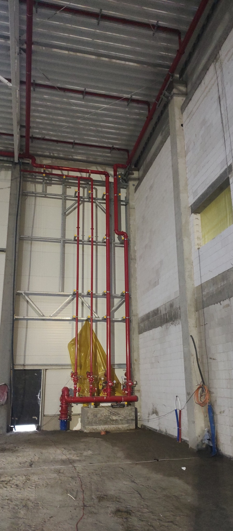 Czerwona instalacja rur przeciwpożarowych w surowym wnętrzu hali, z widocznymi elementami mocującymi i żółtą folią ochronną na dole.