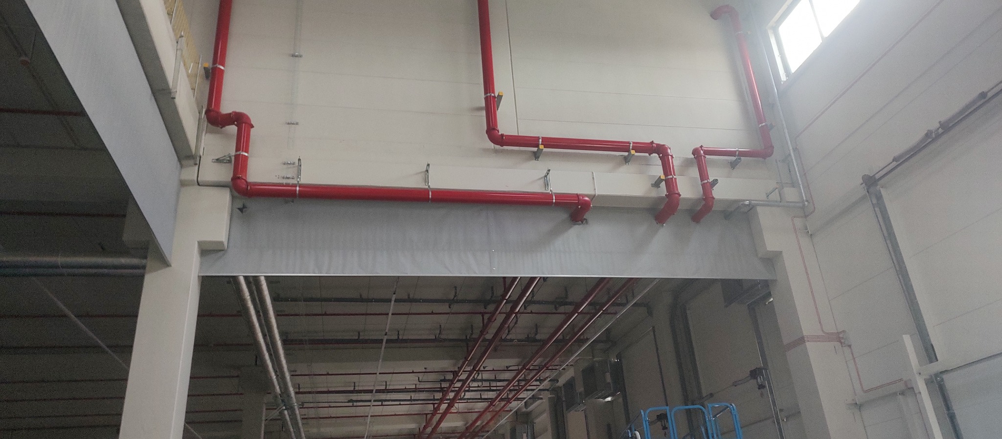 Czerwone rury instalacji przeciwpożarowej zamontowane pod sufitem w hali przemysłowej, widoczne z perspektywy podnośnika.