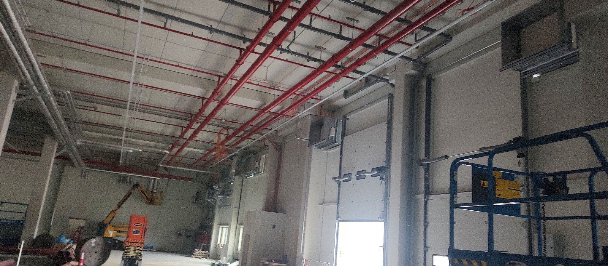 Wnętrze hali przemysłowej w trakcie instalacji rurociągów, widoczne czerwone rury pod sufitem, podnośnik koszowy oraz maszyna budowlana w oddali.