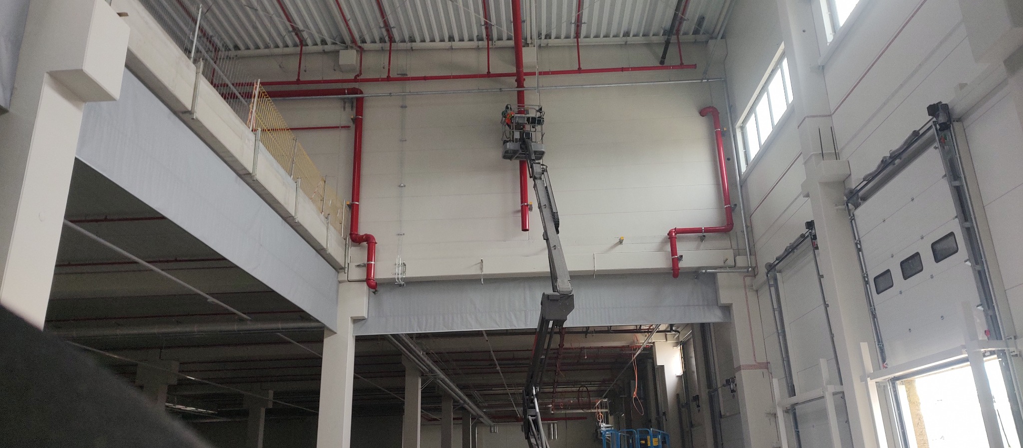 Montaż czerwonej instalacji tryskaczowej w hali przemysłowej z użyciem podnośnika koszowego.