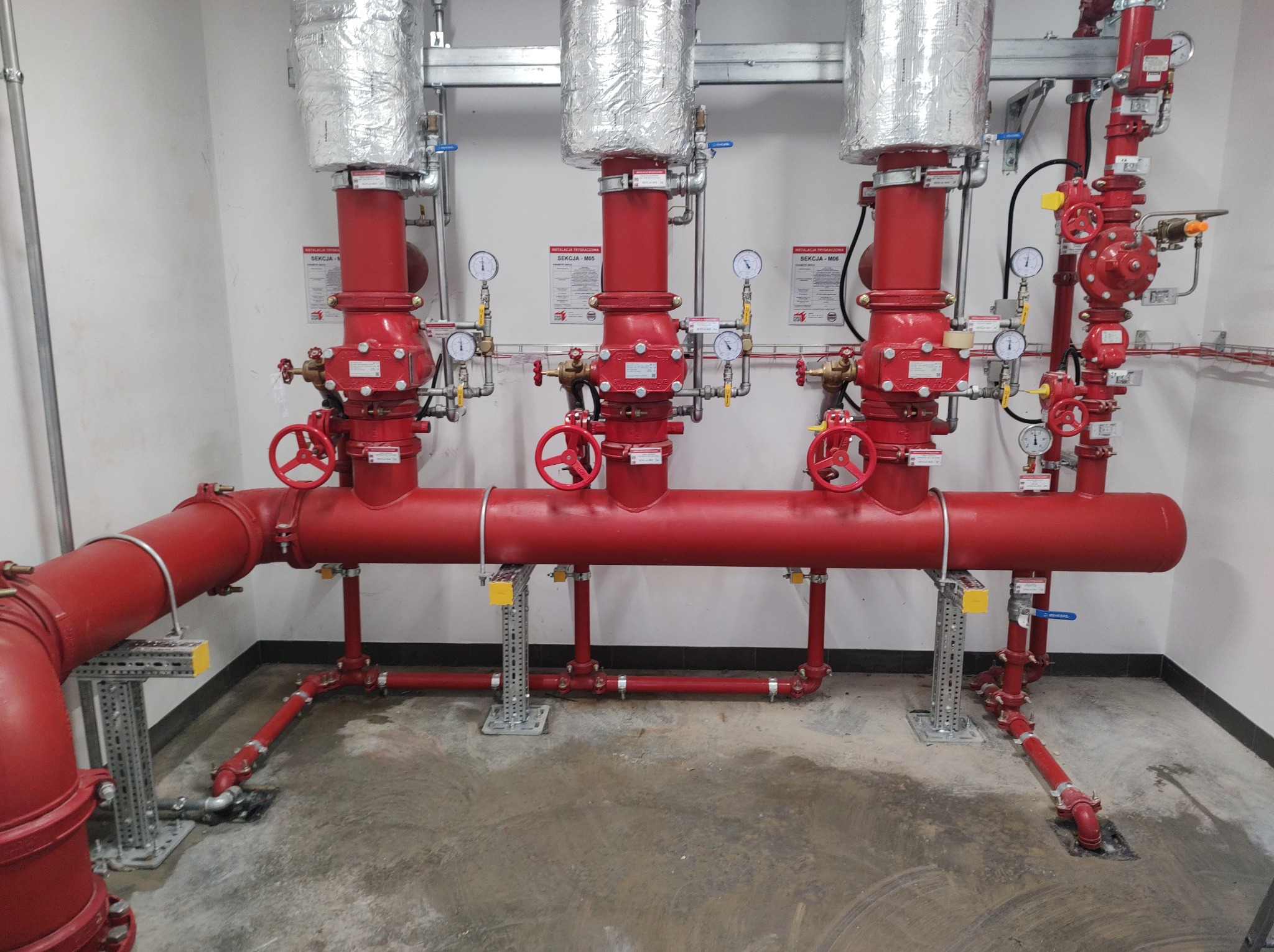 Czerwona instalacja tryskaczowa z manometrami i zaworami, zabezpieczona srebrną folią izolacyjną w pomieszczeniu technicznym.