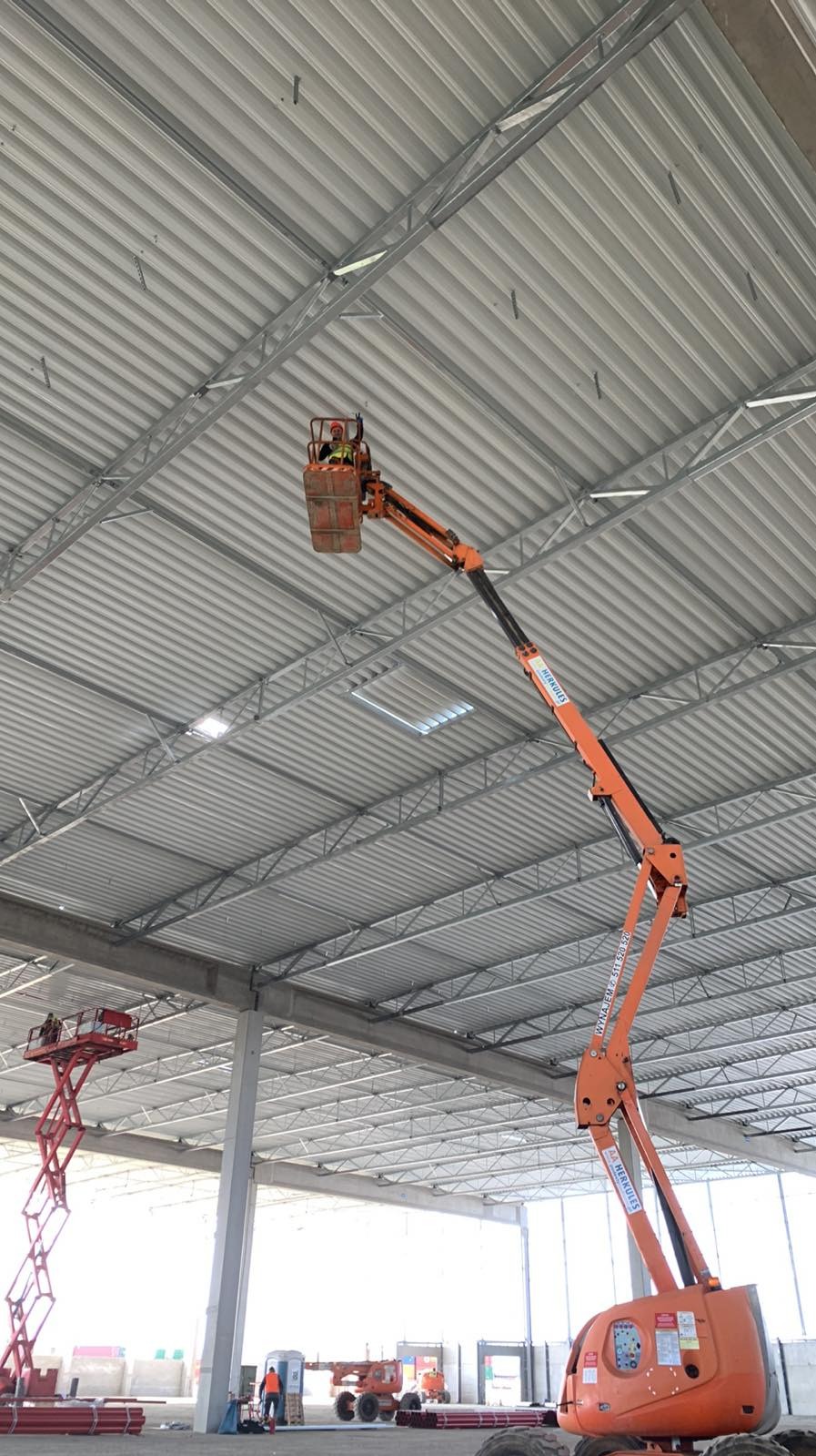 Montaż instalacji pod sufitem hali z użyciem podnośnika koszowego; widoczna kratownica dachu i oświetlenie.