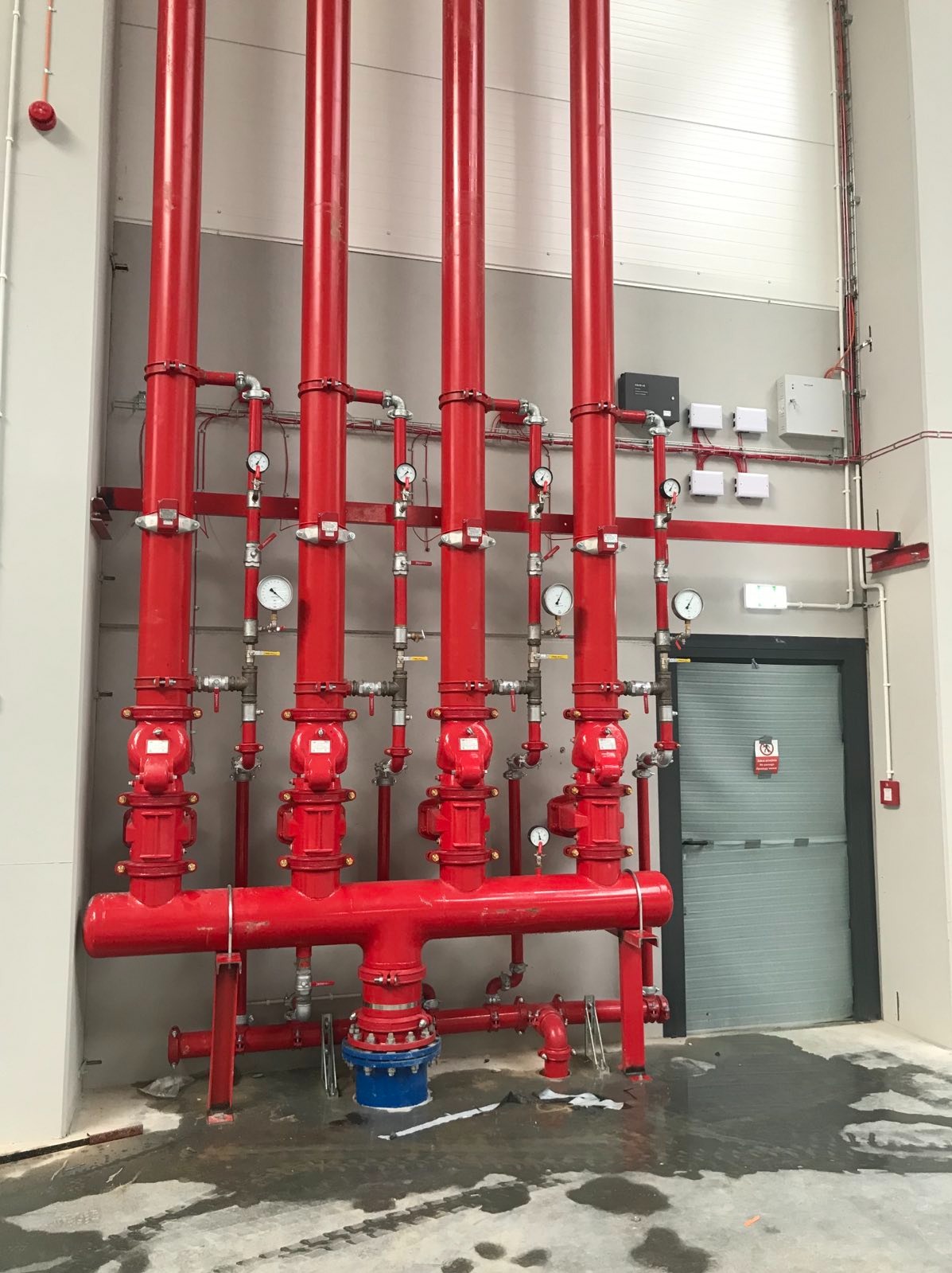 Czerwona instalacja przeciwpożarowa z rurami, zaworami i manometrami w hali przemysłowej, widoczne mokre plamy na betonowej posadzce.