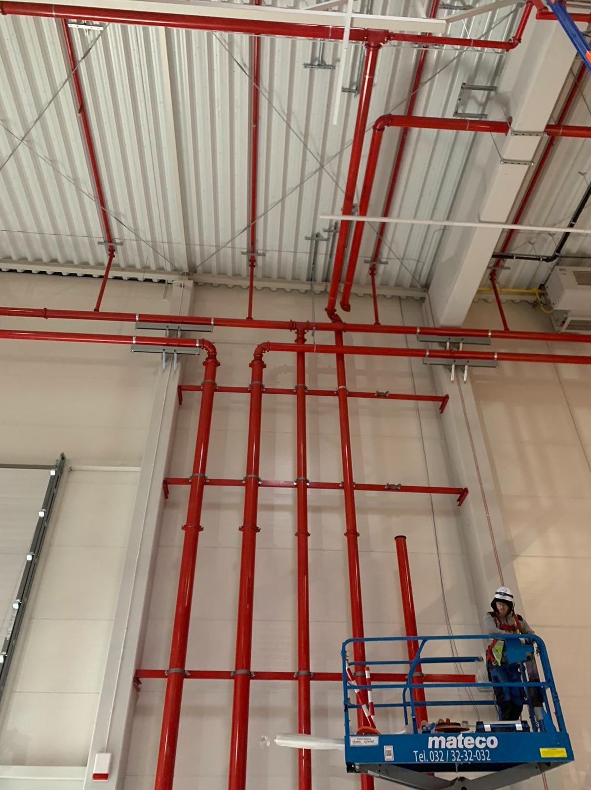 Czerwone rury instalacji przeciwpożarowej zamontowane pod sufitem hali przemysłowej, widoczne mocowania i pracownik na podnośniku koszowym.